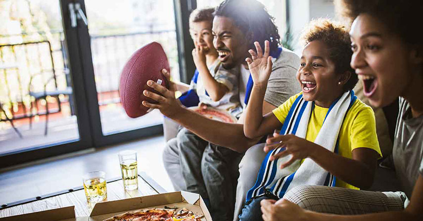 Family watch football, eat pizza with large window and bright sun in background