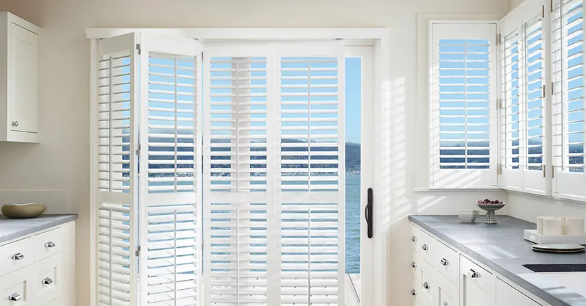 Bifold plantation shutters, a window treatment idea, cover a sliding glass door and windows inside a kitchen.